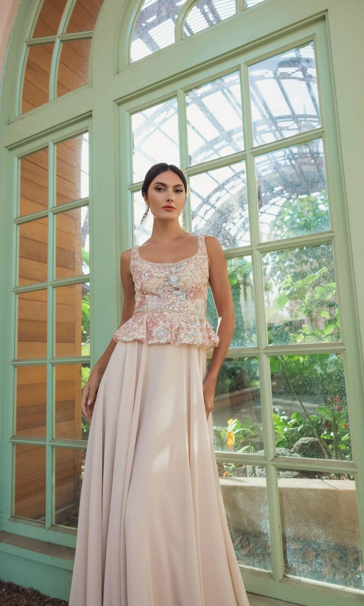 A woman wearing the Mac Duggal 21317 long formal dress in champagne, featuring an A-line chiffon skirt with a floral embroidered bodice and peplum waistline, stands by large green-framed windows in a sunlit conservatory.